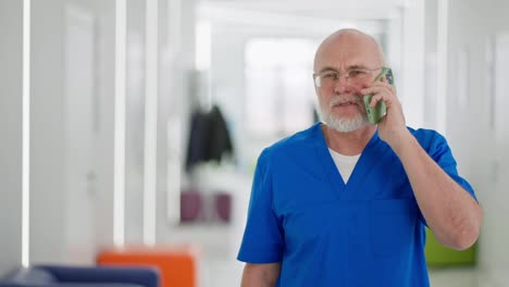 A-confident-man-with-glasses,-an-elderly-doctor-with-a-beard-in-a-blue-uniform-walks-along-the-corridor-of-a-bright-clinic-and-communicates-on-a-green-phone