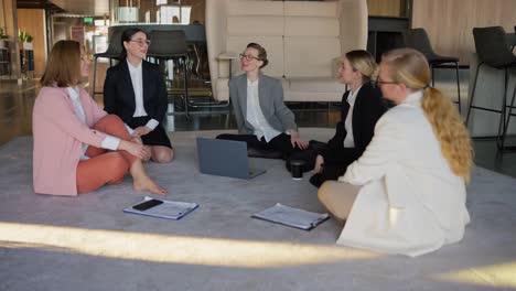 A-group-of-girls-in-business-suits-sit-on-a-gray-carpet-in-the-office-and-communicate-with-each-other-during-a-break-at-work-in-a-modern-office