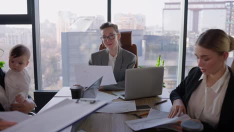 Portrait-of-a-confident-middle-aged-blonde-Business-Woman-wearing-glasses-in-a-gray-suit-while-she-sits-at-an-office-desk-during-a-businesswoman-meeting-in-a-modern-office-with-large-windows