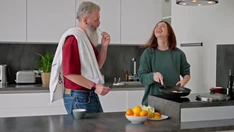 Ein-Glücklicher-älterer-Mann-Mit-Bart-In-Einem-Roten-T-Shirt-Und-Ein-Brünettes-Mädchen-In-Einem-Grünen-Pullover-Tanzen,-Während-Sie-In-Der-Küche-Eines-Modernen-Apartments-Das-Frühstück-Zubereiten