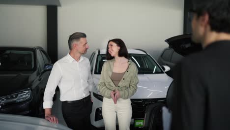 Over-his-shoulder-a-confident-middle-aged-man-with-a-gray-beard-and-his-brunette-wife-in-a-car-showroom-are-talking-with-an-assistant-in-a-suit-about-modern-car-models-and-listening-to-cars-in-a-car-showroom