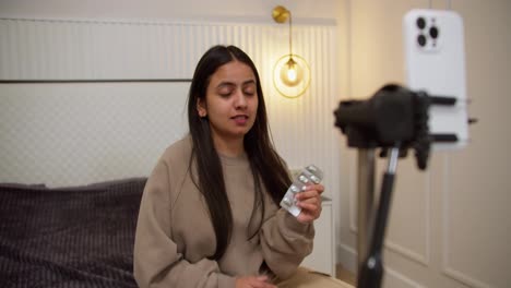 A-confident-brunette-Indian-girl-in-a-gray-jacket-runs-her-video-blog-about-pills-and-talks-about-their-quality-to-her-viewers-using-a-white-smartphone-in-a-modern-apartment