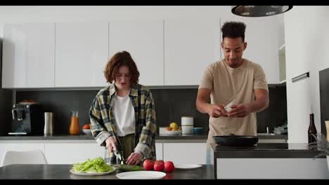 Happy-young-Black-brunette-man-cooking-scrambled-eggs-during-his-breakfast-preparation-along-with-his-mature-young-girlfriend-with-brown-hair-and-bob-hairstyle-in-the-kitchen-in-the-morning