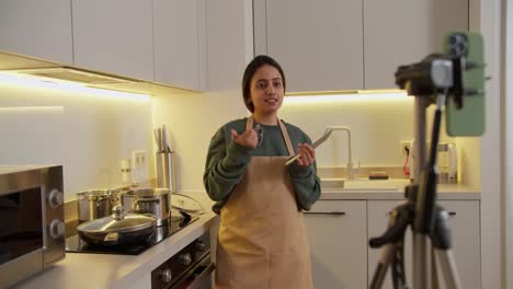 A-happy-brunette-girl-in-a-green-jacket-and-a-beige-apron-runs-a-cooking-and-cooking-blog-in-which-she-talks-about-how-to-properly-prepare-food-and-use-a-kitchen-knife-in-a-modern-apartment-kitchen