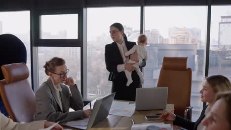 Confident-brunette-businesswoman-with-glasses-in-a-business-suit-holds-a-small-infant-child-in-her-arms-and-communicates-with-her-colleagues-about-an-idea-and-plan-for-work.-During-a-meeting-at-the-table-in-the-office