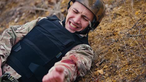 Close-up-a-soldier-in-a-camouflage-uniform-and-body-armor-lying-wounded-in-the-arm-waves-his-hand-to-a-medic-to-provide-first-aid-during-combat-operations-in-the-steppe