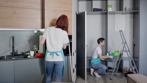 Side-view-of-a-confident-brunette-man-in-a-white-T-shirt-and-blue-apron-cleaning-a-modern-studio-apartment-together-with-his-colleague-a-brunette-girl-while-calling-a-cleaning-company-to-a-modern-apartment