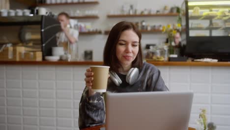 Una-Chica-Morena-Feliz-Con-Una-Chaqueta-Gris-Trabaja-En-Línea-Frente-A-Una-Computadora-Portátil-En-Una-Mesa-En-Un-Café.-Una-Chica-Morena-Trabaja-De-Forma-Remota-Usando-Una-Computadora-Portátil-En-Un-Café