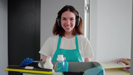 Portrait-of-a-happy-brunette-girl-in-black-wireless-headphones-in-a-white-T-shirt-and-a-blue-apron-who-holds-in-her-hands-a-gray-plastic-basin-with-cleaning-tools-and-detergents-in-a-modern-apartment