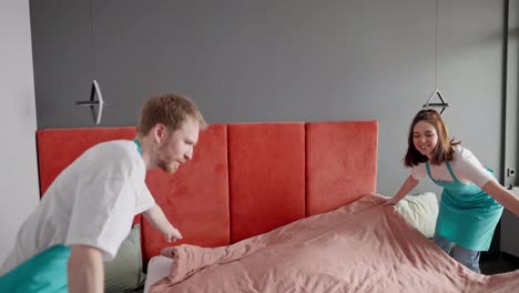 View-from-the-side-of-a-confident-cleaning-guy-a-blond-guy-with-stubble-and-in-a-white-T-shirt-with-a-blue-front-together-with-his-colleague-a-brunette-girl-making-the-bed-in-a-modern-apartment-while-cleaning-on-call