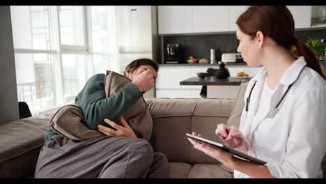 A-tired-and-sick-brunette-man-in-a-green-sweater-hugs-a-pillow-and-sitting-on-the-sofa-tells-a-brunette-girl-to-a-doctor-in-a-white-coat-about-his-symptoms-and-the-girl-writes-them-down-on-her-tablet-during-a-home-examination-in-a-modern-apartment