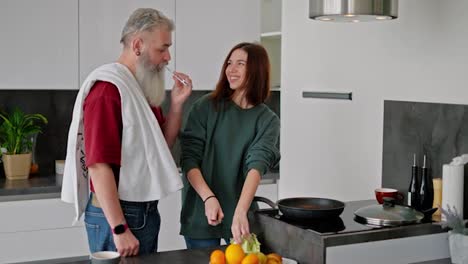 Un-Hombre-Mayor-De-Pelo-Gris-Y-Barba-Frondosa-Con-Una-Camiseta-Roja-Se-Cepilla-Los-Dientes-Mientras-Una-Chica-Morena-Con-Una-Chaqueta-Verde-Prepara-El-Desayuno-Y-Rompe-Huevos-De-Gallina-Para-Hacer-Huevos-Revueltos-En-La-Cocina-De-Un-Apartamento-Moderno.