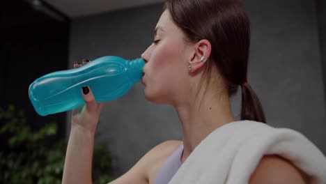 Side-view-close-up-of-a-confident-brunette-girl-in-a-purple-sports-top-with-a-white-towel-on-her-shoulder-drinks-water-from-a-blue-sports-bottle-in-a-modern-apartment-after-playing-sports-in-the-evening