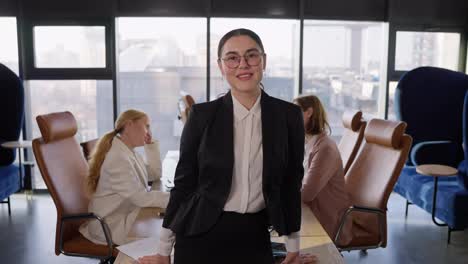 Portrait-of-a-confident-brunette-girl-in-a-business-suit-and-round-glasses-posing-near-a-wooden-table-and-her-colleagues-in-an-office-with-large-windows