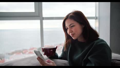 Happy-and-busy-brunette-girl-in-a-dark-green-sweater-typing-on-her-mobile-phone-and-holding-a-brown-mug-of-tea-in-a-modern-apartment-while-sitting-on-the-sofa