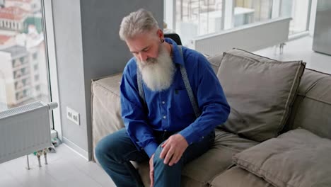 An-elderly-man-with-gray-hair-and-a-bushy-beard-massages-his-knee-while-sitting-on-a-brown-sofa-in-a-modern-apartment.-An-elderly-man-stretches-his-knee-after-a-long-walk-in-a-modern-apartment-with-large-windows