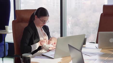 A-confident-brunette-girl-in-round-glasses-in-a-business-uniform-sits-at-a-table-in-front-of-a-laptop-in-the-office-and-breastfeeds-her-baby-without-being-distracted-from-work-in-a-modern-office