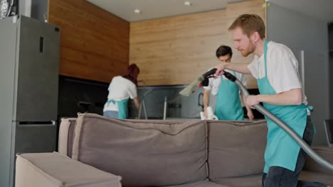 Confident-male-blond-cleaner-in-a-white-T-shirt-and-blue-apron-cleans-a-brown-sofa-using-a-vacuum-cleaner-with-his-colleagues-in-a-modern-studio-apartment