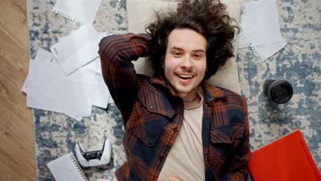 Portrait-of-a-happy-brunette-guy-in-a-plaid-shirt-who-lies-on-a-pillow-on-the-floor-among-sheets-of-papers-coffee-laptop-and-joystick-at-home