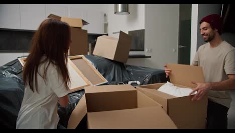 A-happy-brunette-man-with-stubble-in-a-beige-T-shirt-brings-things-in-a-special-box-for-his-brunette-girlfriend-in-a-white-T-shirt-in-a-new-apartment-they-are-sorting-things-out-of-the-boxes-and-settling-in-after-moving