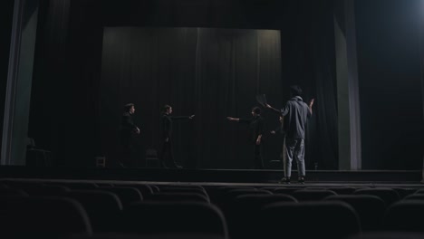 A-group-of-young-actors-in-black-suits-rehearse-and-expressively-show-their-roles-together-with-their-director-on-stage-during-preparation-for-a-performance-in-the-theater