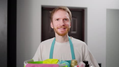 Portrait-of-a-confident-blond-guy-in-a-blue-apron-with-a-large-gray-plastic-basin-with-cleaning-tools-in-a-modern-apartment