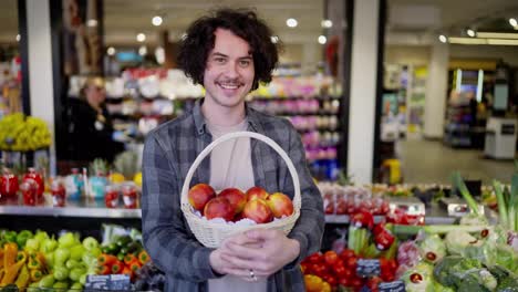 Portrait-of-a-happy-brunette-guy-with-a-mustache-holding-a-basket-of-red-peaches-in-his-hands-and-posing-in-a-grocery-store