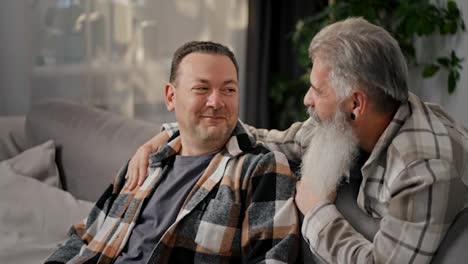 Happy-elderly-man-with-gray-hair-and-a-lush-beard-communicates-and-hugs-his-brunette-boyfriend-with-stubble-in-a-plaid-shirt-on-a-gray-sofa-in-a-modern-apartment