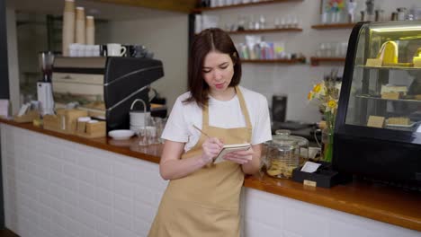 Portrait-of-a-happy-girl-waitress-in-a-yellow-apron-and-white-T-shirt-who-makes-notes-in-her-notebook-regarding-orders-in-a-cafe