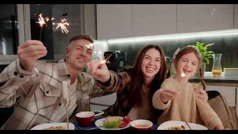 Portrait-of-a-happy-brunette-woman-in-a-plaid-shirt-together-with-her-husband-and-little-daughter-with-a-braided-hairstyle-who-are-holding-sparklers-in-their-hands-having-fun-and-rejoicing-while-sitting-at-the-dining-table-during-a-festive-dinner-in-the-evening-in-a-modern-apartment