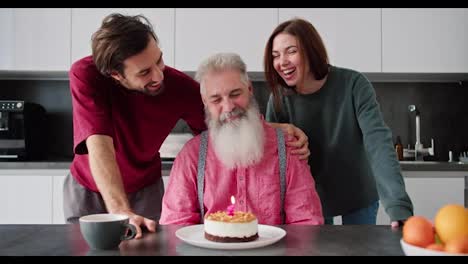 Ein-Glücklicher-älterer-Mann-Mit-Grauem-Haar-Und-üppigem-Bart-In-Einem-Rosa-Hemd-Bläst-Eine-Kerze-Auf-Einem-Kuchen-Aus-Und-Seine-Erwachsenen-Kinder,-Ein-Mann-Und-Ein-Brünettes-Mädchen-In-Einer-Grünen-Jacke-Gratulieren-Ihrem-Vater-Zu-Seinem-Geburtstag-In-Einer-Modernen-Wohnung