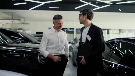 A-confident-guy-a-brunette-assistant-in-glasses-and-a-black-body-suit,-communicates-with-a-middle-aged-man-in-a-white-shirt-about-a-specific-model-of-a-modern-white-car-in-a-car-showroom