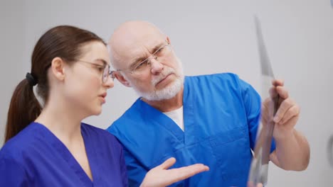 A-confident-elderly-man-an-experienced-doctor-in-a-blue-uniform,-communicates-with-his-colleague-a-brunette-girl-in-round-glasses-about-chest-fluorography-in-a-modern-clinic