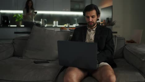 Confident-brunette-man-with-stubble-in-a-jacket-and-beige-shorts-works-using-a-gray-laptop-while-sitting-on-a-sofa-in-a-modern-apartment-in-the-evening