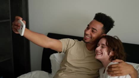 A-happy-Black-skinned-brunette-man-in-a-cream-T-shirt-lies-on-the-bed-with-his-young-adult-brunette-girlfriend-with-a-bob-hairstyle-in-a-white-T-shirt-and-takes-a-selfie-using-a-white-phone