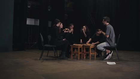 View-from-Zoom-in-of-a-confident-group-of-young-actors-in-black-uniforms-sitting-around-a-pizza-and-taking-a-slice-during-their-lunch-and-break-while-preparing-for-a-performance-in-the-theater