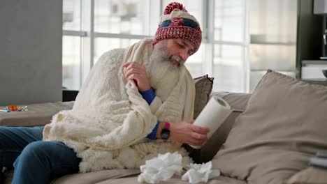 An-elderly-man-with-a-thick-gray-beard-in-a-red-cap-wearing-a-white-plaid-and-blowing-his-nose-in-a-napkin-while-he-is-sick-and-has-a-runny-nose-and-sits-at-home-on-a-modern-sofa-in-the-apartment