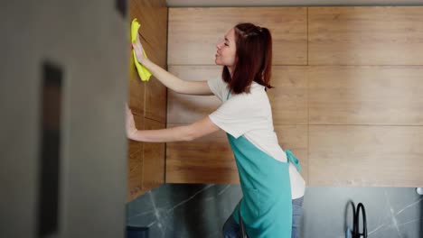 Side-view-of-a-confident-brunette-cleaning-lady-girl-in-a-white-t-shirt-and-blue-apron-wiping-the-upper-cabinets-in-a-modern-kitchen-with-a-wooden-texture-using-a-yellow-rag-while-cleaning-a-studio-apartment-on-call