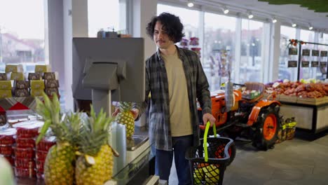 Ein-Mann-Mit-Lockigem-Haar-Und-Hemd-Wiegt-Ananas-Auf-Einer-Waage-In-Einem-Supermarkt,-Während-Er-In-Einem-Lebensmittelgeschäft-Einkauft