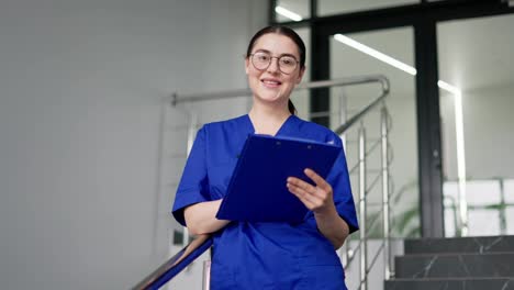 Porträt-Einer-Selbstbewussten-Und-Glücklichen-Brünetten-Ärztin-Mit-Runder-Brille-In-Herbstuniform,-Die-Eine-Blaue-Tablette-In-Den-Händen-Hält,-Lächelt-Und-Posiert-In-Einer-Modernen-Klinik-Auf-Der-Treppe