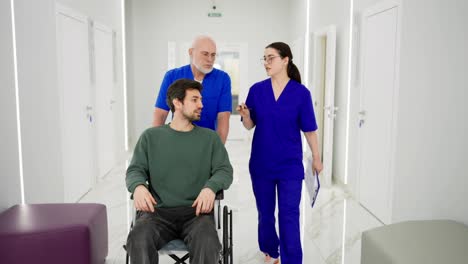 A-confident-elderly-doctor-in-glasses-with-a-gray-beard-and-in-a-blue-uniform-takes-a-man-to-wheelchairs-for-the-disabled-while-a-brunette-girl-talks-with-the-doctor-about-the-rehabilitation-of-a-young-brunette-guy-in-a-green-shirt-after-a-broken-leg