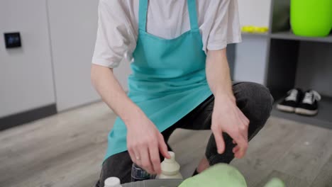 Close-up-portrait-of-a-confident-guy-a-blond-cleaner-in-a-white-T-shirt-and-a-blue-apron-who-put-detergents-and-cleaning-tools-in-a-gray-plastic-basin-while-cleaning-in-a-modern-apartment