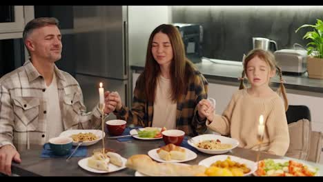 Familia-Feliz,-Mujer-Morena-Con-Una-Camisa-A-Cuadros-Junto-Con-Su-Esposo-De-Mediana-Edad-Con-Cabello-Gris-Y-Su-Pequeña-Hija-Rezando-Y-Dando-Gracias-Por-La-Comida-Antes-De-Comenzar-Una-Cena-En-Una-Cocina-Moderna