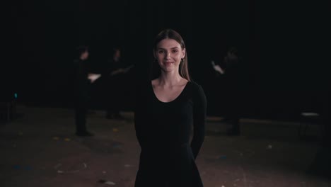 Portrait-of-a-confident-young-girl-actor-in-a-black-suit-among-the-actors-who-are-preparing-for-a-performance-and-rehearsing-on-stage-in-a-theater-with-black-curtains