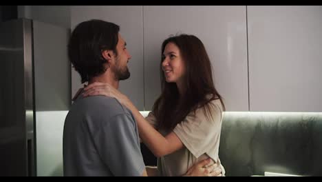 A-happy-brunette-girl-in-a-beige-T-shirt-hugs-her-brunette-boyfriend-with-stubble-in-a-blue-T-shirt-in-the-evening-in-a-modern-kitchen.-Happy-couple-guy-and-girl-hugging-during-their-romantic-evening-in-the-kitchen