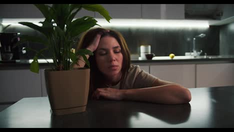 A-tired-brunette-girl-in-a-beige-t-shirt-leans-on-a-black-table-and-holds-her-head-while-thinking-near-an-indoor-flower-of-a-green-plant-in-the-kitchen-in-the-evening-portrait