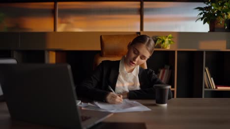 A-confident-blonde-businesswoman-in-a-black-jacket-and-business-uniform-sits-at-a-table-and-writes-her-ideas-on-important-sheets-of-paper-in-front-of-a-laptop-in-a-sunny-office