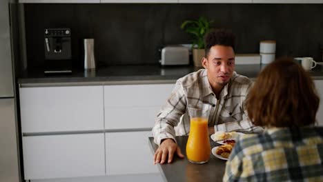 Over-the-shoulder-a-happy-young-guy-with-Black-skin-color-a-brunette-in-a-checkered-beige-shirt-communicates-with-his-girlfriend-with-brown-hair-and-a-bob-hairstyle-during-breakfast-in-a-modern-kitchen