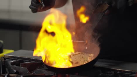 Nahaufnahme:-Ein-Professioneller-Koch-In-Schwarzer-Uniform-Gießt-Alkohol-In-Eine-Heiße-Bratpfanne-Mit-Einem-Gericht,-Um-Feuer-Zu-Erzeugen,-Und-Verwendet-Die-Flambiertechnik,-Während-Er-In-Einer-Restaurantküche-Ein-Gericht-Zubereitet.-Der-Koch-Gibt-Alkohol-In-Die-Bratpfanne-Und-Erzeugt-Ein-Kontrolliertes,-Schönes,-Helles-Feuer-In-Der-Küche