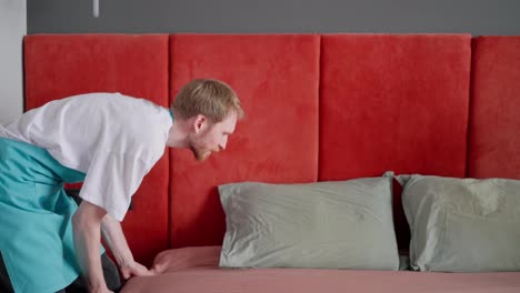 Side-view-of-a-blond-guy-with-a-beard-a-cleaner-in-a-white-T-shirt-and-a-blue-apron-together-with-his-colleague-a-brunette-guy-makes-the-bed-neatly-in-the-bedroom-cleaning-company